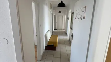 Langer, heller Flur mit weißen Wänden, Fliesenboden und einer gelben Sitzbank.