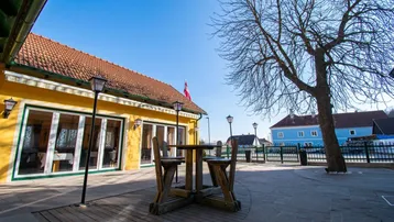 Außenbereich eines gelben Gebäudes mit rotem Ziegeldach, einer Holzterrasse und einem gepflasterten Sitzbereich unter blauem Himmel.
