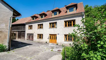 Traditionelles Wohnhaus mit mehreren Fenstern und einem gepflasterten Vorplatz unter blauem Himmel.