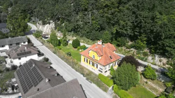 Luftaufnahme eines charmanten gelben Hauses mit rotem Dach, umgeben von üppiger Natur und einer Straße.