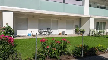 Neubau-Gebäude mit gepflegtem Rasen, Blumenbeeten und einer Terrasse im Erdgeschoss.