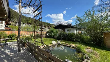 Gepflegter Garten mit Teich und Terrasse, umgeben von grüner Natur und Bergblick.
