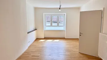 Helles Zimmer mit Parkettboden, weißen Wänden und einem Fenster, das Tageslicht hereinlässt.