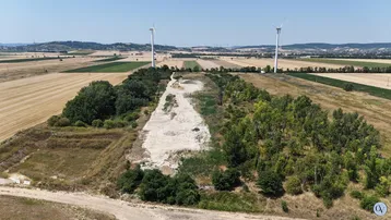 Luftaufnahme einer weiten Landschaft mit Feldern, Bäumen und Windrädern am Horizont unter blauem Himmel.
