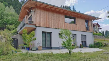 Modernes Haus mit Holzfassade und Balkon in grüner, hügeliger Umgebung.