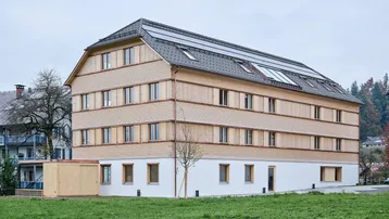 Modernes Mehrfamilienhaus mit Holzfassade, Satteldach und integrierten Solarpaneelen auf dem Dach.