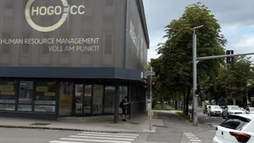 Gebäudeansicht an einer Straßenecke mit Zebrastreifen und umliegender Vegetation unter bewölktem Himmel.