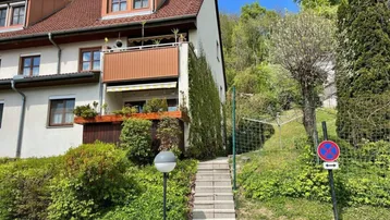 Mehrfamilienhaus mit mehreren Balkonen und begrüntem Hang im Hintergrund unter blauem Himmel.