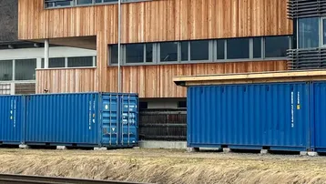 Blaue Schiffscontainer stehen vor einem modernen Gebäude mit Holzfassade und großen Fenstern, im Hintergrund sind Berge sichtbar.