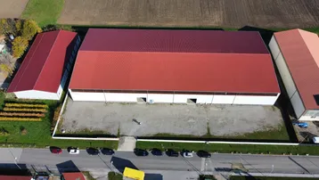 Luftaufnahme eines großen Industriegebäudes mit rotem Dach, umgeben von Feldern und einer Straße mit parkenden Autos.