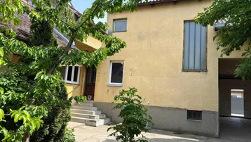 Fassade eines älteren Gebäudes mit gelbem Putz und verschiedenen Fenstern, umgeben von Bäumen.