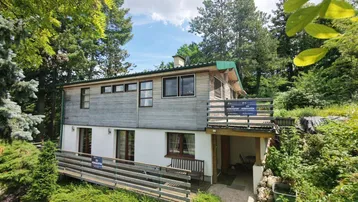 Rustikales Holzhaus umgeben von üppigem Grün und hohen Bäumen, mit Balkon und Terrasse.