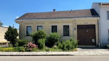 Historisches Gebäude mit beigefarbener Fassade, Satteldach und einem kleinen Vorgarten mit Blumenbeeten und Sträuchern.