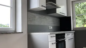 Moderne Küche mit grauen und weißen Schränken, integriertem Backofen und Dunstabzugshaube, hell durch Fenster.