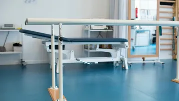Heller Therapieraum mit Parallelbarren, Untersuchungsliege und Spiegel für Rehabilitationsübungen.