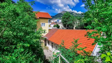 Haus mit rotem Ziegeldach, umgeben von üppigem Grün und mit Bergblick unter blauem Himmel.