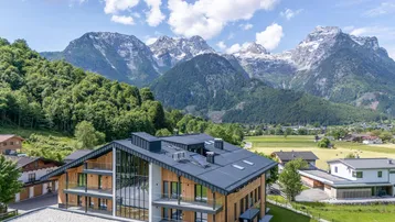 Modernes Gebäude mit Holzfassade und dunklem Dach, eingebettet in eine grüne Landschaft mit majestätischen Bergen im Hintergrund.