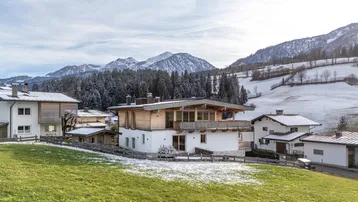 Modernes Haus in alpiner Landschaft mit schneebedeckten Bergen im Hintergrund und grünen Wiesen.