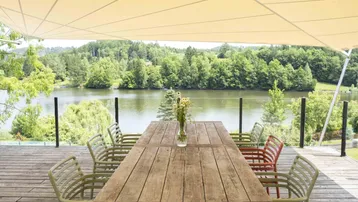 Große Holzterrasse mit Esstisch und Stühlen, überdacht von einem Sonnensegel, mit Blick auf den See.