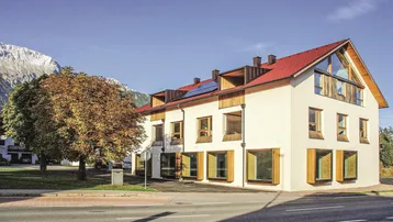 Modernes Gebäude mit weißer Fassade, rotem Dach und großen Fenstern vor alpiner Bergkulisse