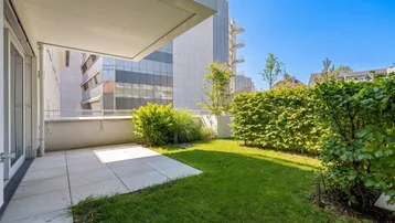 Grüne Gartenfläche mit gepflegtem Rasen und Hecke, angrenzend an eine geflieste Terrasse.