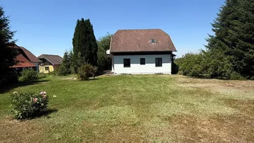 Kleines Haus mit braunem Dach und weißer Fassade, umgeben von einem großen Garten mit Bäumen.