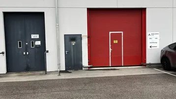 Außenansicht eines Industriegebäudes mit mehreren Laderampen und Türen, sowie einem Parkplatz davor.