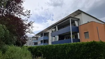 Modernes Mehrfamilienhaus mit blauen Balkonen und orangefarbener Fassade, umgeben von grünen Hecken.