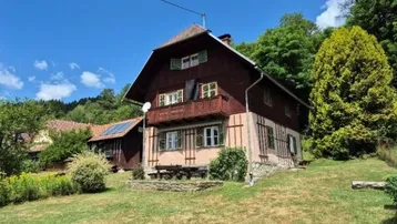 Haus im Landhausstil mit Holzbalkon und grüner Umgebung unter blauem Himmel.