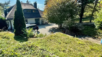 Großzügige Auffahrt zum Haus mit viel Grün und Baumbestand unter blauem Himmel.