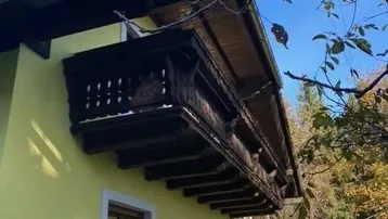 Traditioneller Holzbalkon an einem Haus mit gelber Fassade unter blauem Himmel und Bäumen.