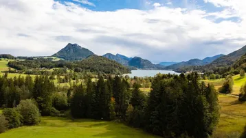 Malerische Landschaft mit weiten grünen Feldern, einem See und majestätischen Bergen unter blauem Himmel.