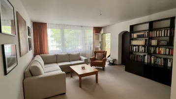 Helles Wohnzimmer mit beigem Ecksofa, Teppichboden und großem Bücherregal.