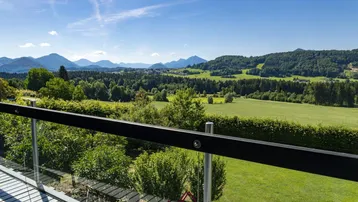 Blick vom Balkon auf eine weite grüne Landschaft mit Wäldern und Bergen unter blauem Himmel.