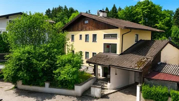 Gelbes Einfamilienhaus mit braunem Ziegeldach, umgeben von viel Grün und Bäumen, inklusive Carport.