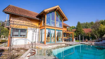 Großzügiges Einfamilienhaus mit Holzfassade, großem Pool und Gartenbereich unter blauem Himmel.