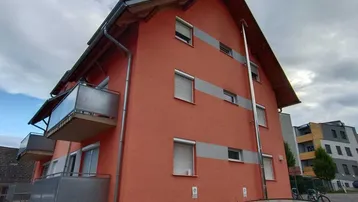 Gebäudeansicht mit orangefarbener Fassade, Balkonen und Rollläden unter bewölktem Himmel.