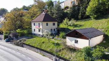 Luftaufnahme eines freistehenden Hauses mit Nebengebäude und Hanglage an einer Straße.