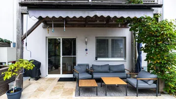Gepflasterte Terrasse mit bequemen Gartenmöbeln, Markise und Zugang zum Innenbereich.