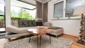 Gemütliches Wohnzimmer mit beigem Sofa, Holztisch und großen Fenstern mit Blick ins Grüne.