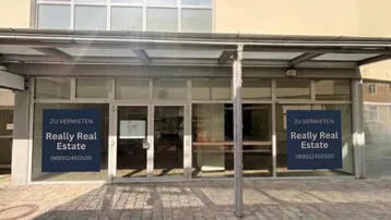 Außenansicht eines Geschäftsgebäudes mit großen Glasfronten und "Zu Vermieten"-Schildern an den Fenstern.