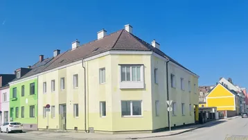Gebäudeansicht von der Straßenseite mit gelber Fassade und mehreren Fenstern in einem städtischen Umfeld.