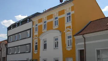 Ansicht eines Gebäudes mit gelber und weißer Fassade sowie parkenden Autos davor.
