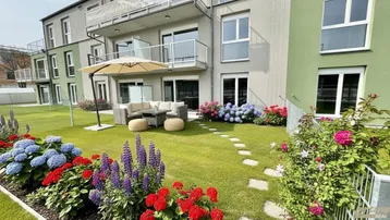 Gepflegter Gartenbereich mit möblierter Terrasse und blühenden Blumenbeeten vor dem modernen Gebäude.