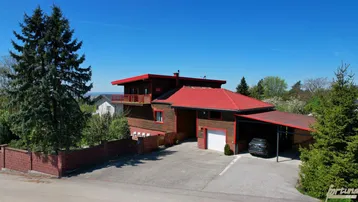 Großzügiges Holzhaus mit rotem Dach, Carport und weitläufiger Auffahrt in ländlicher Umgebung.