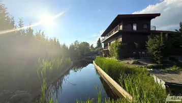 Dunkles Holzhaus mit Balkonen am Wasser, umgeben von üppiger Natur unter strahlendem Sonnenschein.