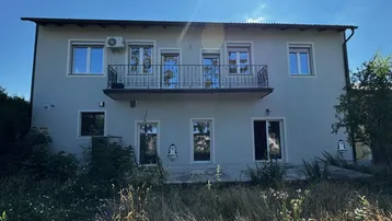Graue Hausfassade mit Balkon und mehreren Fenstern, umgeben von wilder Vegetation.