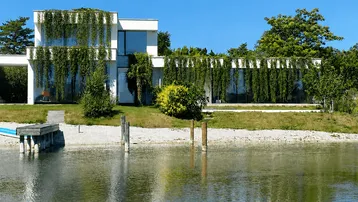 Moderne Villa am Seeufer mit üppiger Begrünung und privatem Steg unter blauem Himmel.
