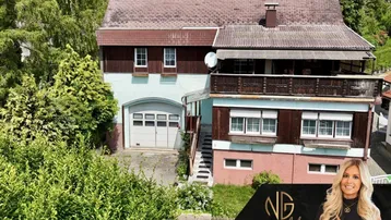 Älteres Mehrfamilienhaus mit Satteldach, Balkon und integrierter Garage, umgeben von viel Grün.