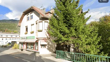 Gebäude mit Ladenlokal im Erdgeschoss und Wohnungen darüber, umgeben von Bäumen und Bergen.